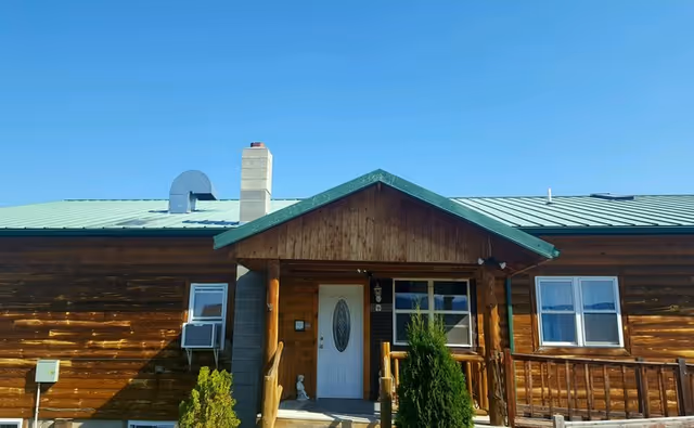 Single-story wooden building front with a green metal roof, covered porch, white door and shrubs.