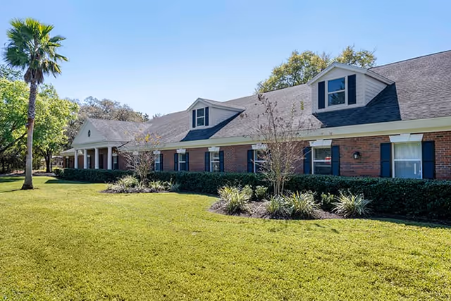 Exterior view of a single-story brick building with a dark shingled roof, dormer windows, and blue shutters. The building is surrounded by a well-maintained lawn, small shrubs, and a palm tree under a clear blue sky.