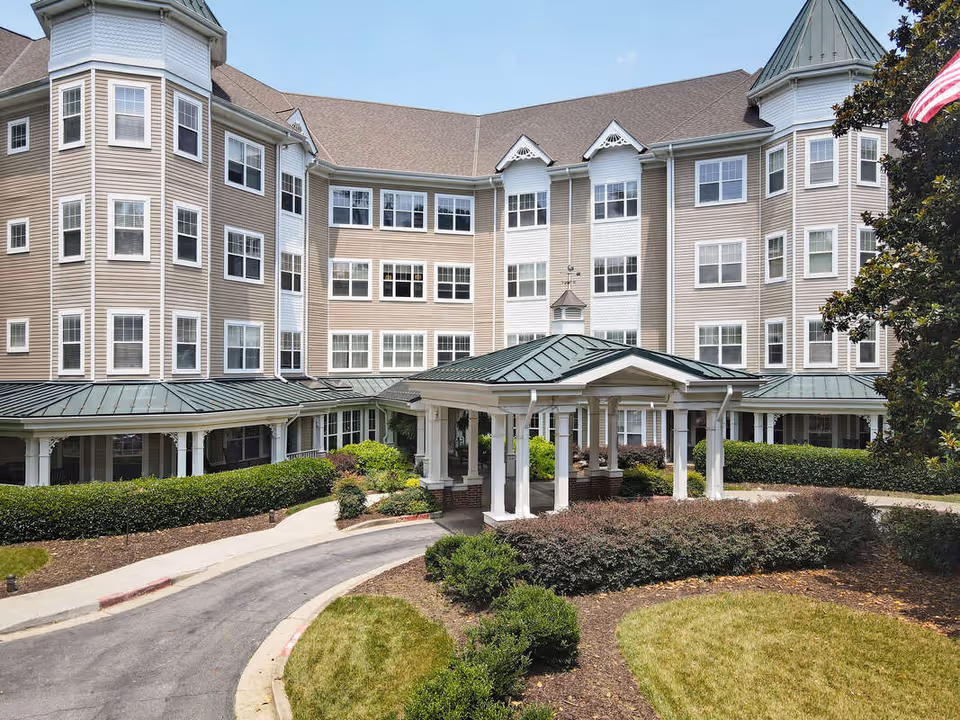 Exterior view of Sunrise at Huntcliff Summit II, a multi-story senior living facility with beige siding, numerous windows, a covered entrance with white columns, and landscaped grounds including bushes and a lawn.