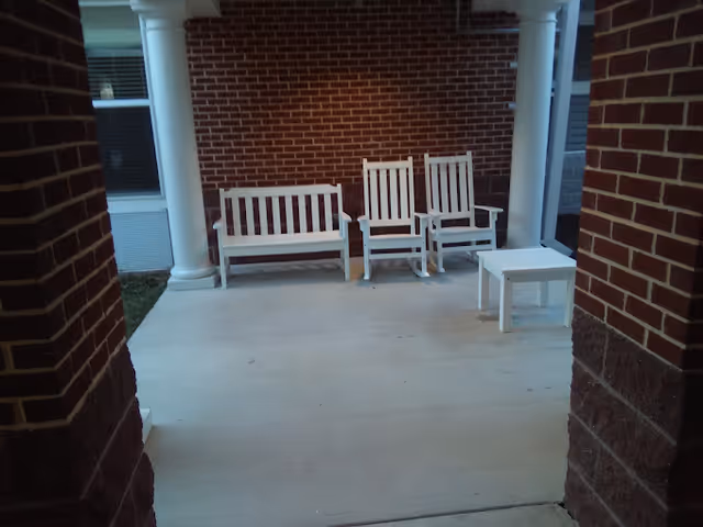 A covered outdoor patio area with white wooden furniture including a bench, two rocking chairs, and a small table. The patio is framed by brick columns and walls, with a window visible on the left side.