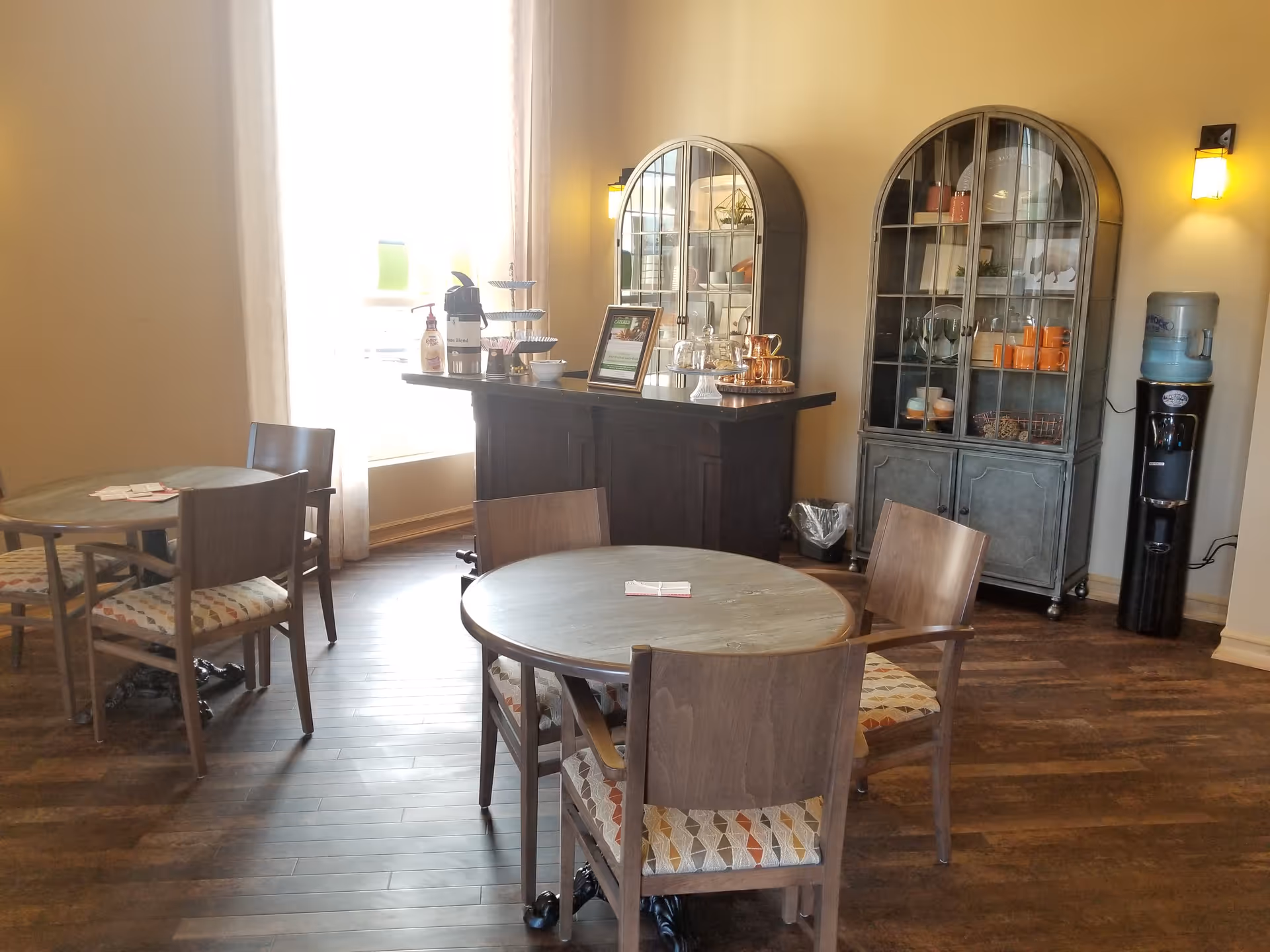 A cozy dining area with two round wooden tables, each surrounded by four wooden chairs with patterned cushions. In the background, there is a dark wooden buffet table with coffee and snacks, flanked by two glass-front cabinets displaying cups and decorative items. A water cooler stands to the right, and soft wall sconces provide warm lighting. A large window with sheer curtains allows natural light to fill the room.