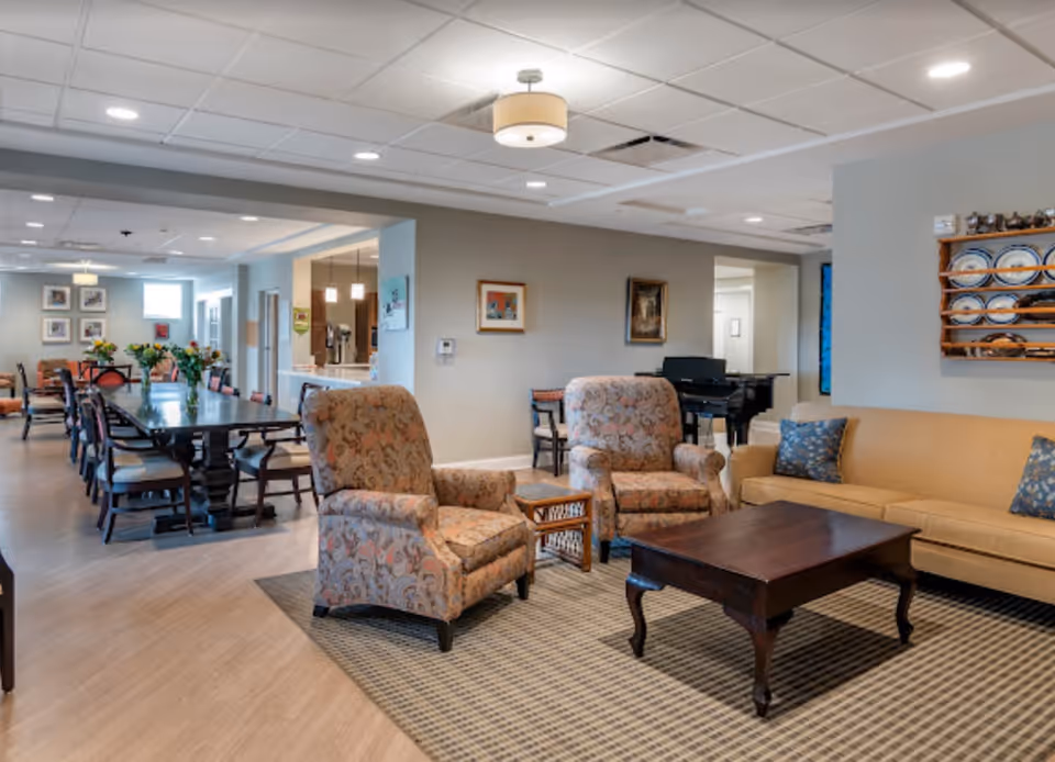 Community lounge with upholstered armchairs, a sofa and coffee table next to a long dining table in a senior living facility.