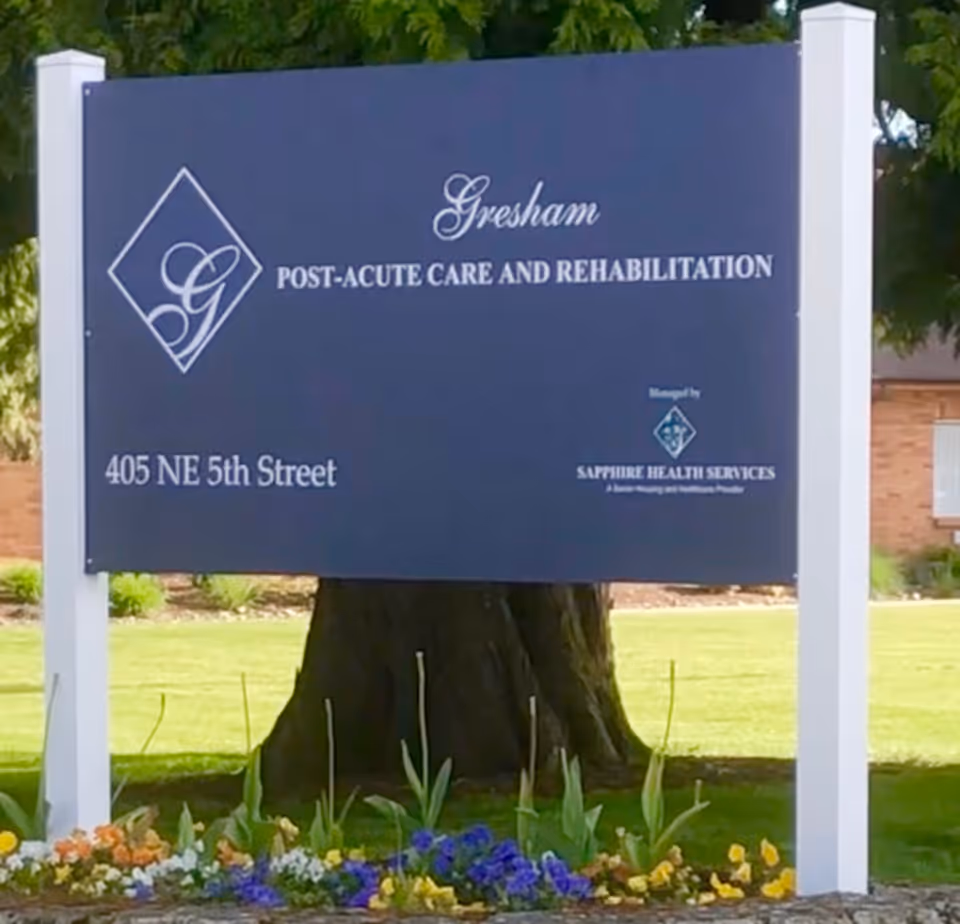 Outdoor sign for Gresham Post-Acute Care and Rehabilitation with the address '405 NE 5th Street' mounted between white posts in front of a tree and flowerbed.