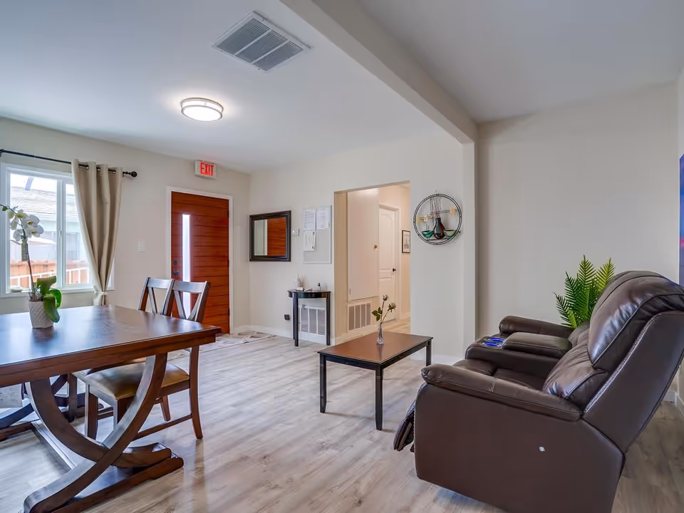 A bright and clean living room and dining area with light wood flooring. The dining area has a wooden table with two chairs and a potted orchid on the table. The living area features two brown leather recliners, a small coffee table with a single flower in a vase, and a green plant in the corner. There is a wooden front door with a vertical window panel, a window with beige curtains, and a small table with a mirror and some documents on the wall.