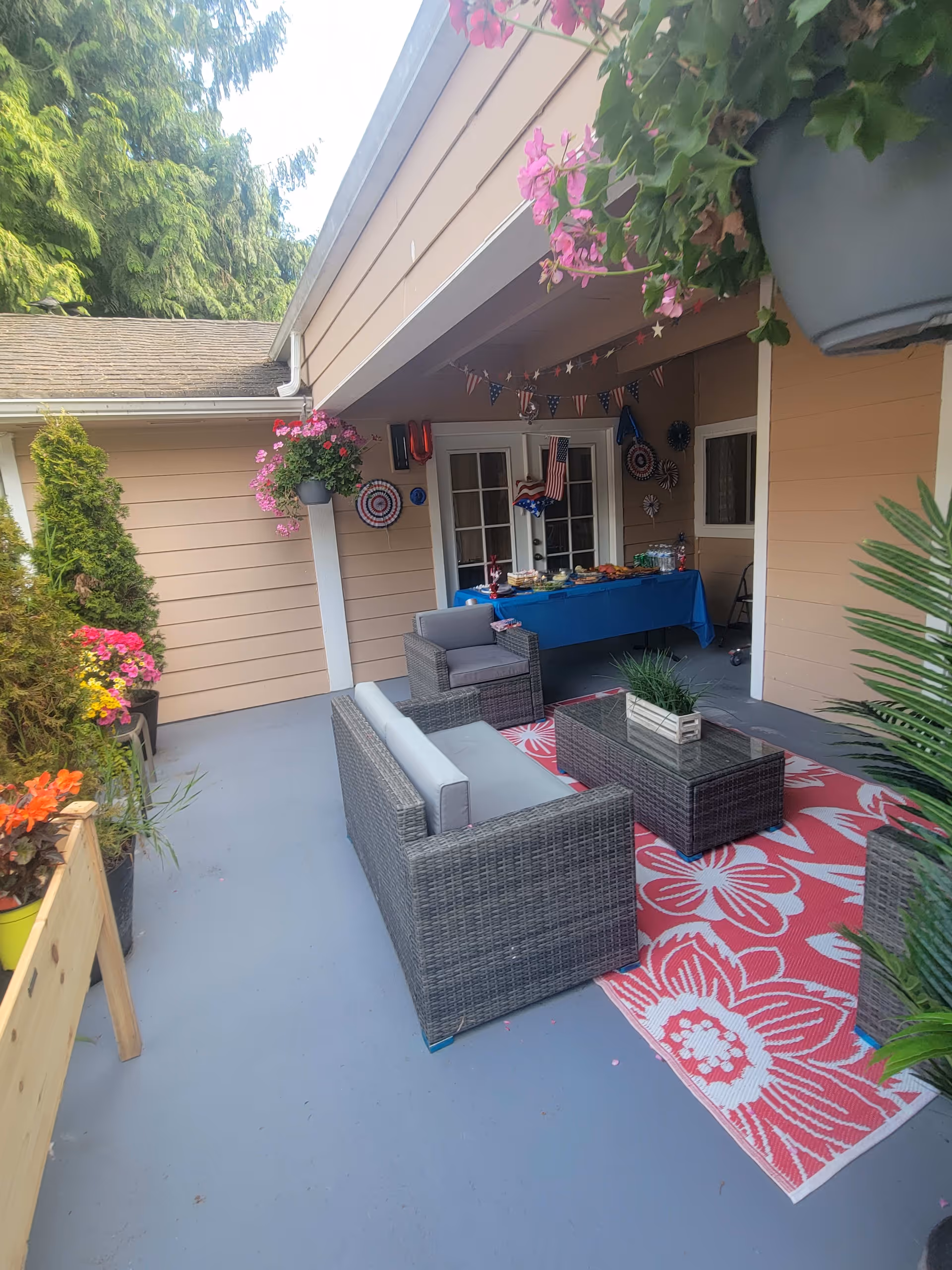 Outdoor covered patio area with wicker furniture including a sofa, armchair, and glass-top coffee table on a red and white floral rug. There are hanging flower pots with pink flowers, potted plants along the side, and a table covered with a blue tablecloth set up with food and drinks. The area is decorated with patriotic bunting and decorations.