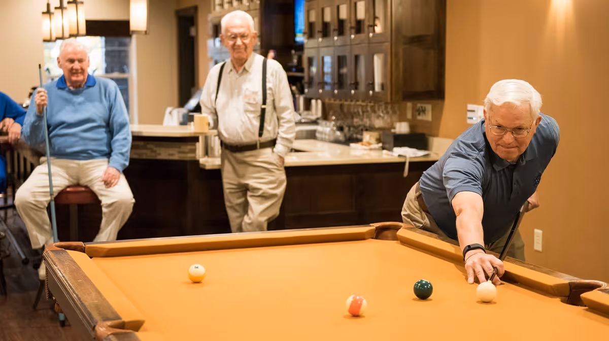Three elderly men in a recreational room with a pool table. One man is leaning over the table aiming to hit the cue ball with a pool cue, while the other two men watch. The room has warm lighting and wooden cabinetry in the background.