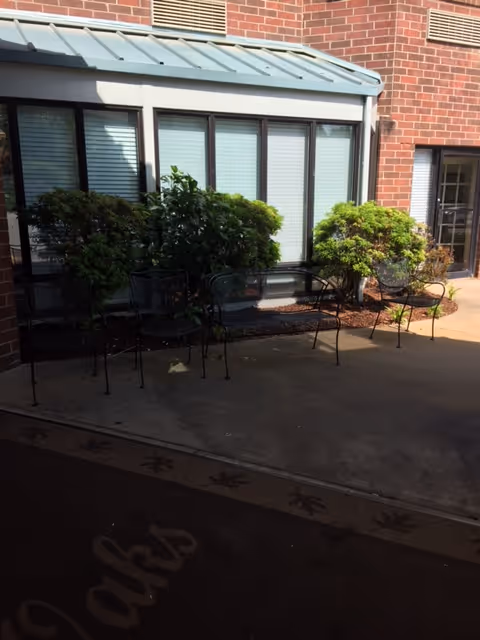 Outdoor seating area with black metal chairs and a bench placed on a concrete patio next to a brick building with windows and some green bushes.