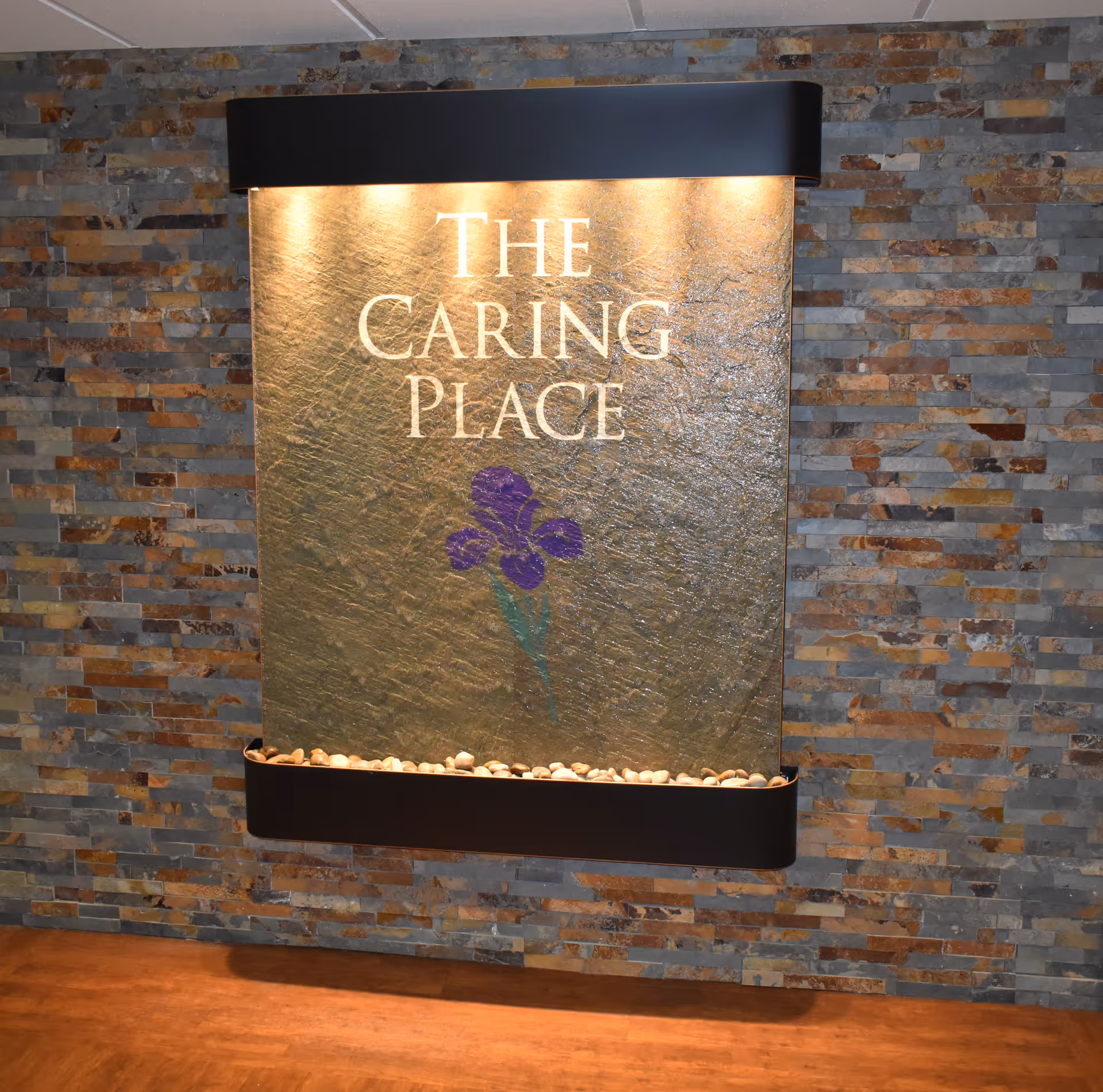 Illuminated wall water feature sign reading "The Caring Place" with a purple flower mounted on a stone-tile interior wall.