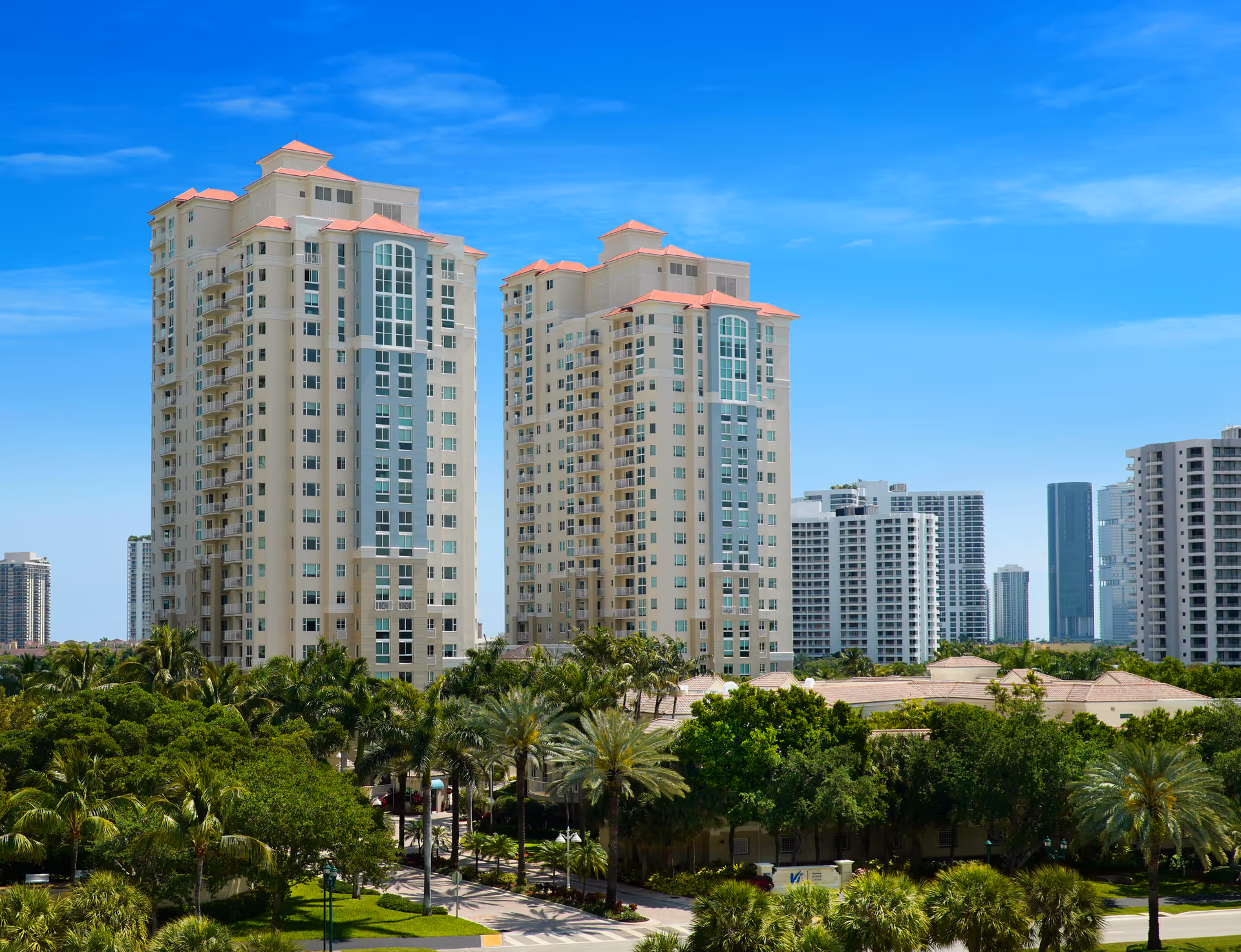 Two tall residential buildings with beige walls and red roofs surrounded by lush green trees and palm trees under a clear blue sky in an urban area.