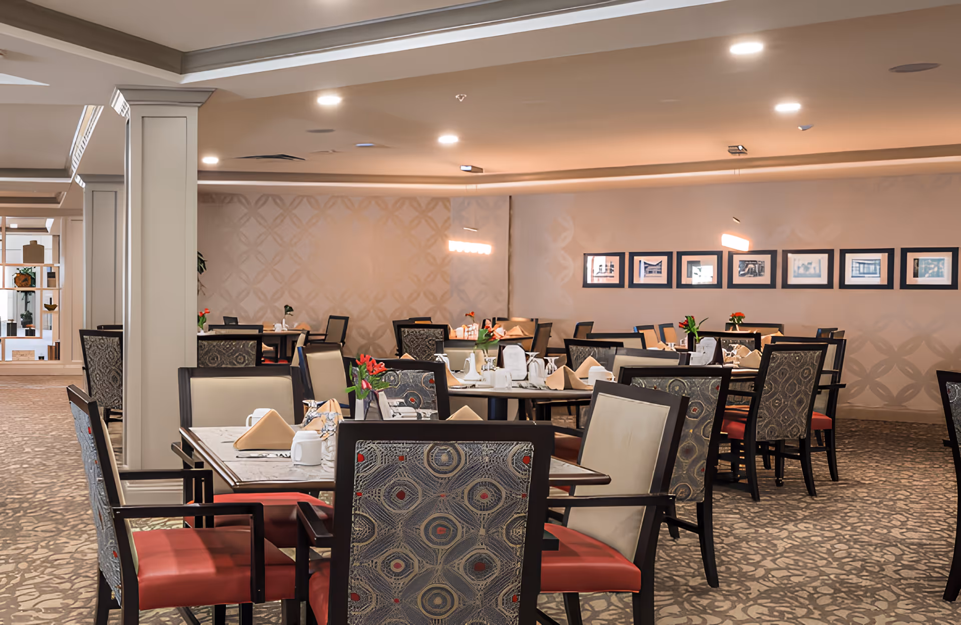 A spacious dining room with multiple tables set with white cups, napkins, and small flower vases. The chairs have patterned backs and red seats. The room has soft lighting, a patterned carpet, and framed pictures on the far wall.