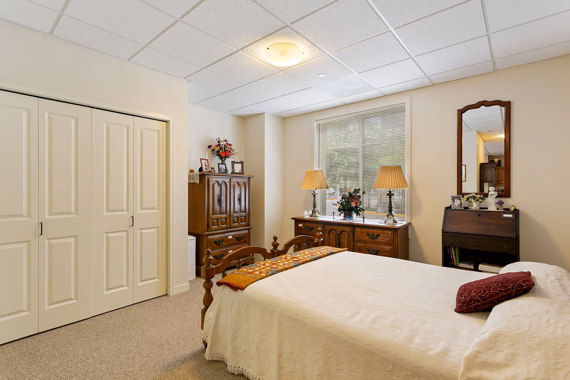 A cozy bedroom with a bed covered in a cream-colored quilt and a red pillow. The room features wooden furniture including a dresser with two lamps and floral decorations, a cabinet with framed photos and flowers, and a small desk with a mirror above it. There is a window with blinds letting in natural light and a closet with sliding doors.
