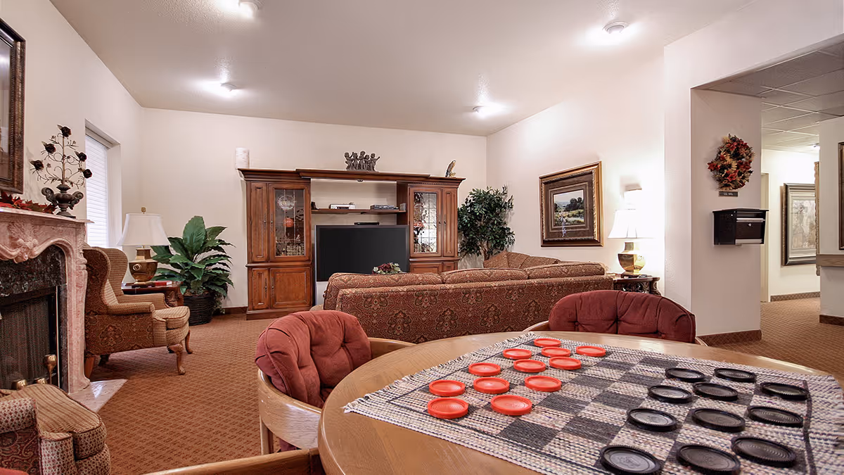 A cozy living room area in a senior living facility with patterned sofas, a wooden entertainment center with a TV, a fireplace with decorative items, and a round table with a checkers game set up. The room is warmly lit with ceiling lights and table lamps, and decorated with plants and framed artwork.