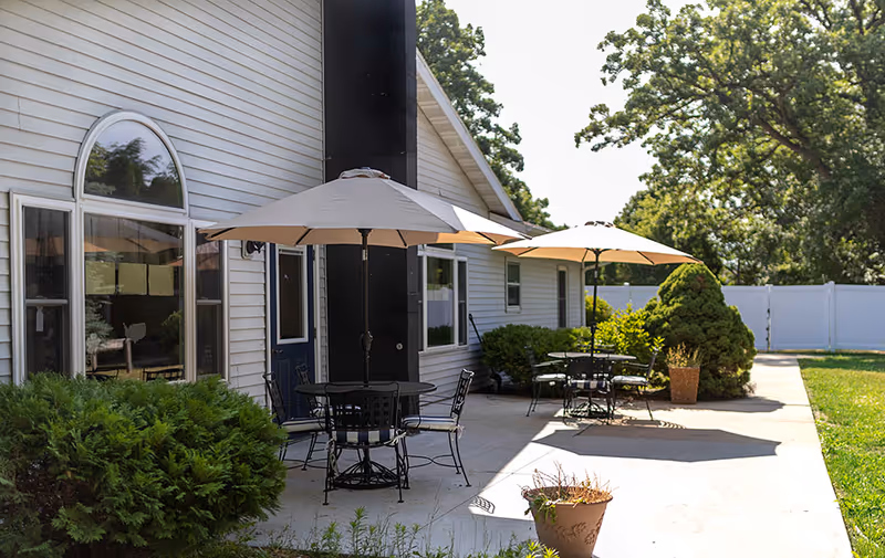 Outdoor patio with metal tables, chairs, and umbrellas beside a light-colored building and landscaped yard.
