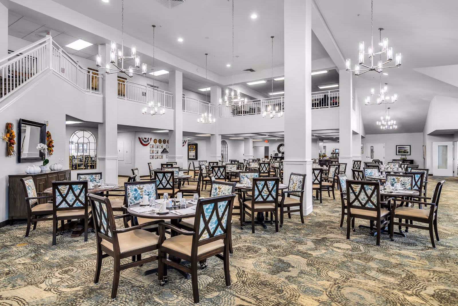 Spacious dining room in a senior living facility with multiple round tables and chairs arranged neatly. The chairs have patterned backs and beige seats. The room features high ceilings with modern chandeliers, white walls, and a patterned carpet. There is a staircase and a balcony area visible on the left side, and some wall decorations including a mirror and framed pictures.