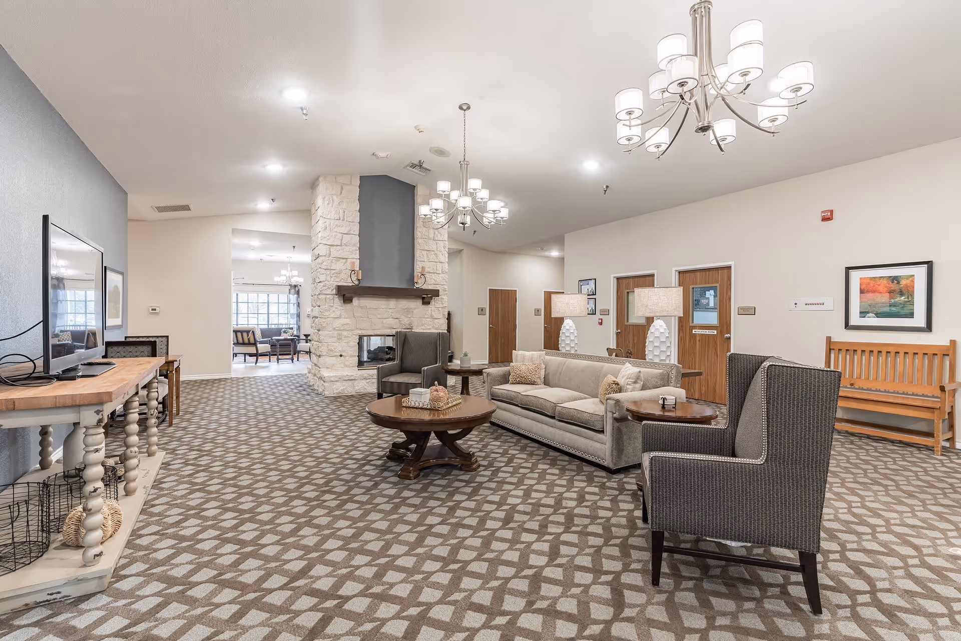 A spacious and well-lit living room area in an assisted living facility featuring a patterned carpet, a stone fireplace with a wooden mantle, a beige sofa with decorative pillows, two armchairs, round wooden coffee tables, two table lamps, a wooden bench, framed artwork on the walls, and a flat-screen TV on a wooden console table.