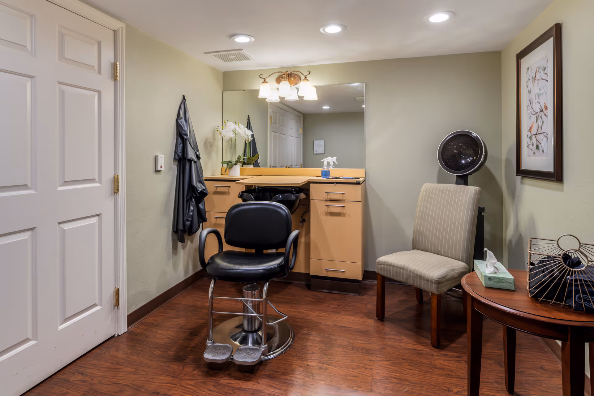 Small salon/styling area with a barber chair facing a mirror, countertop cabinets, a waiting chair and side table.