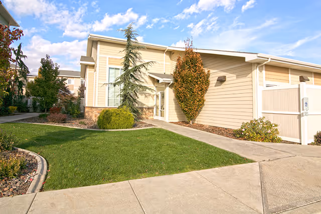 Exterior view of a single-story beige building with a sloped roof, surrounded by a well-maintained lawn, shrubs, and trees under a partly cloudy blue sky.