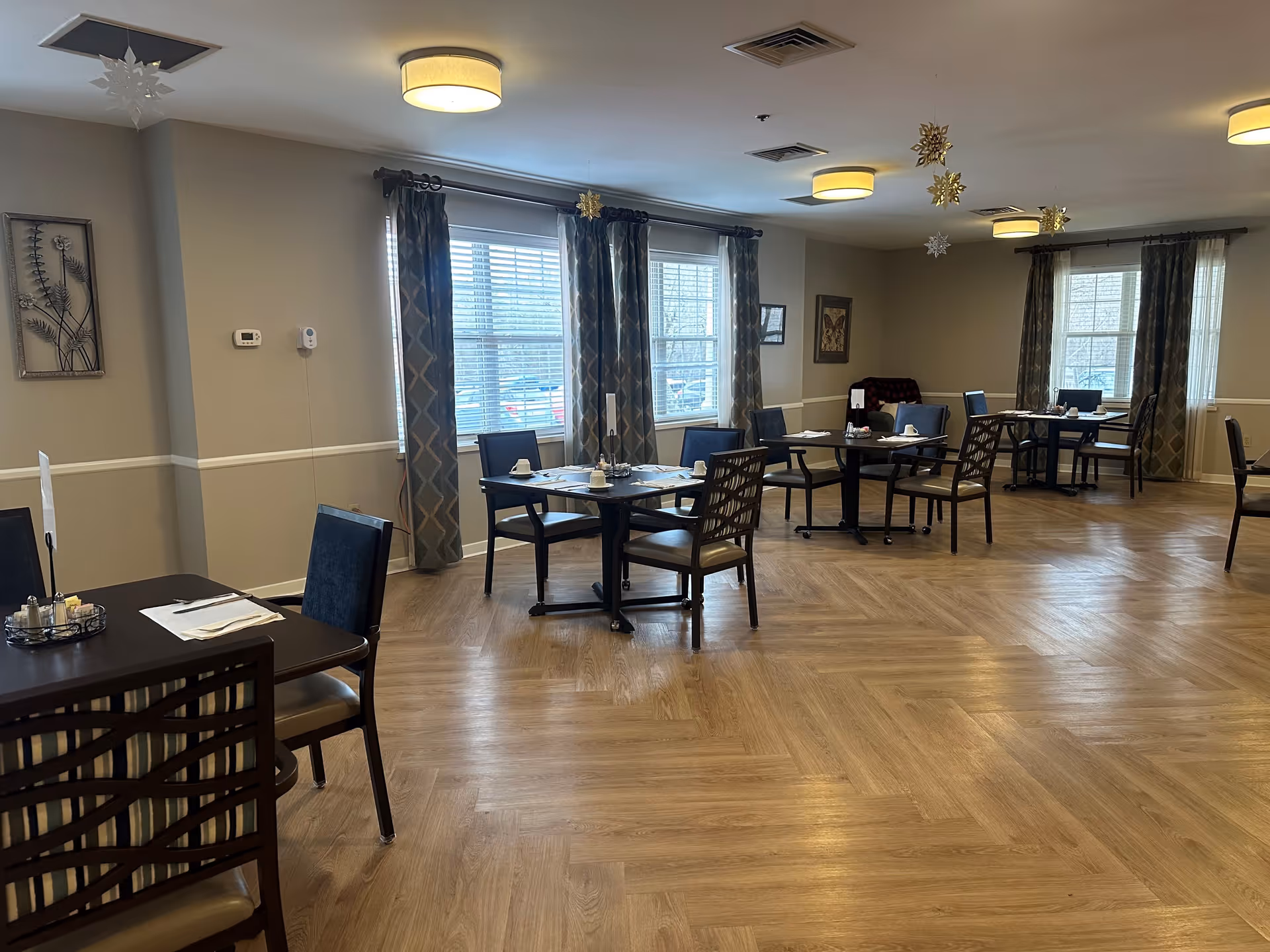 A dining room with several tables and chairs arranged neatly. Each table is set with napkins, cups, and condiments. The room has large windows with patterned curtains, light wood flooring, and ceiling lights. Decorative snowflakes hang from the ceiling, and framed artwork is visible on the walls.