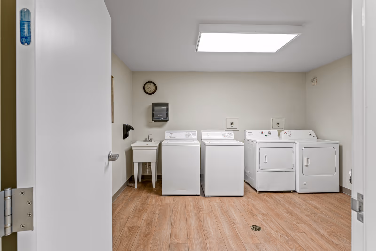 Laundry room with two washing machines and two dryers lined up against a beige wall. There is a small utility sink with a faucet to the left of the machines, a wall-mounted paper towel dispenser above the sink, and a clock on the wall. The floor has a wood-like finish and the ceiling has a large rectangular light panel.