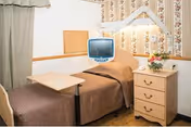 Small patient bedroom with a single bed, overbed table, bedside dresser with lamp and flowers, and a mounted TV.