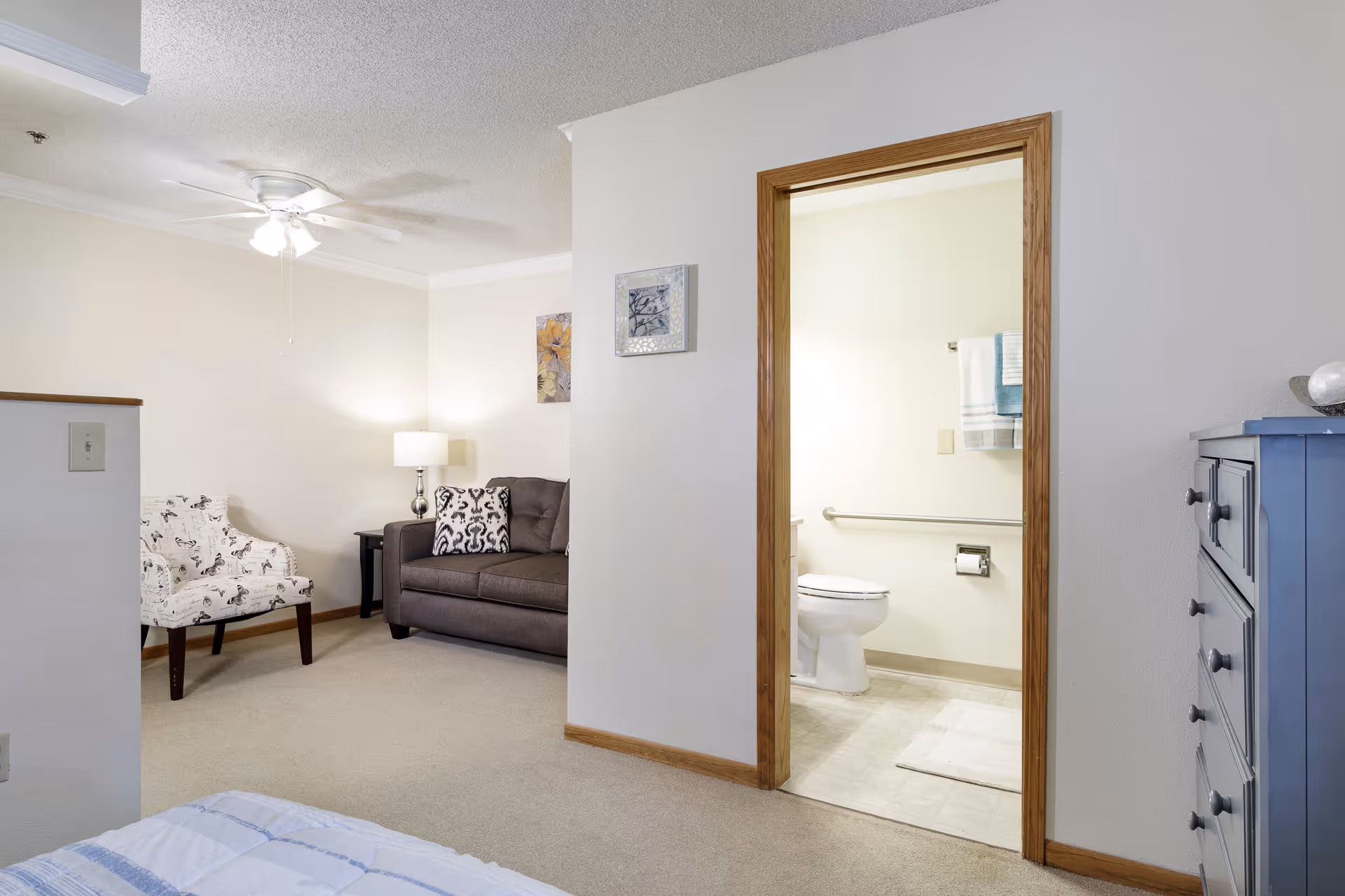 View of a living area with a gray couch, a patterned armchair, a side table with a lamp, and a ceiling fan. To the right, there is an open doorway leading to a bathroom with a toilet, grab bar, and towels hanging on the wall. A blue dresser is partially visible on the right side of the image.