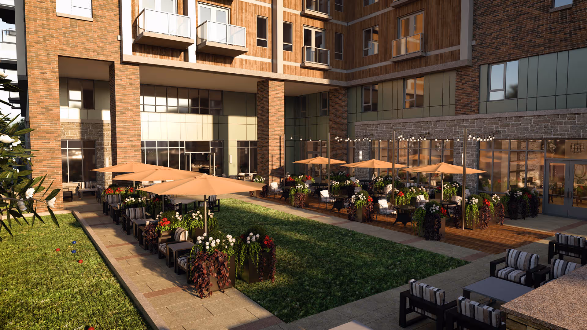 Outdoor courtyard area of a senior living facility with green grass, multiple seating areas with cushioned chairs, tables, and large beige umbrellas. The building has large windows and balconies overlooking the courtyard. There are decorative plants and string lights adding ambiance to the space.