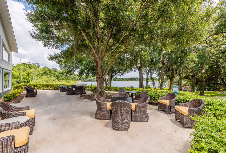 Outdoor patio area with multiple wicker chairs with yellow cushions arranged around small tables under large trees. The patio overlooks a lake with greenery and trees surrounding the area. Part of a building is visible on the left side of the image.