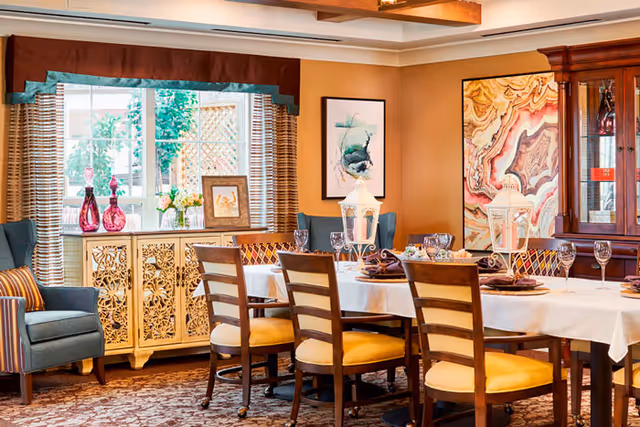 A warmly decorated dining room with a long table set for a meal, featuring wooden chairs with yellow cushions, white tablecloth, glassware, and decorative lanterns. The room includes a patterned carpet, a blue armchair with striped pillows, a carved wooden sideboard with vases and flowers, framed artwork on the walls, and a wooden china cabinet.