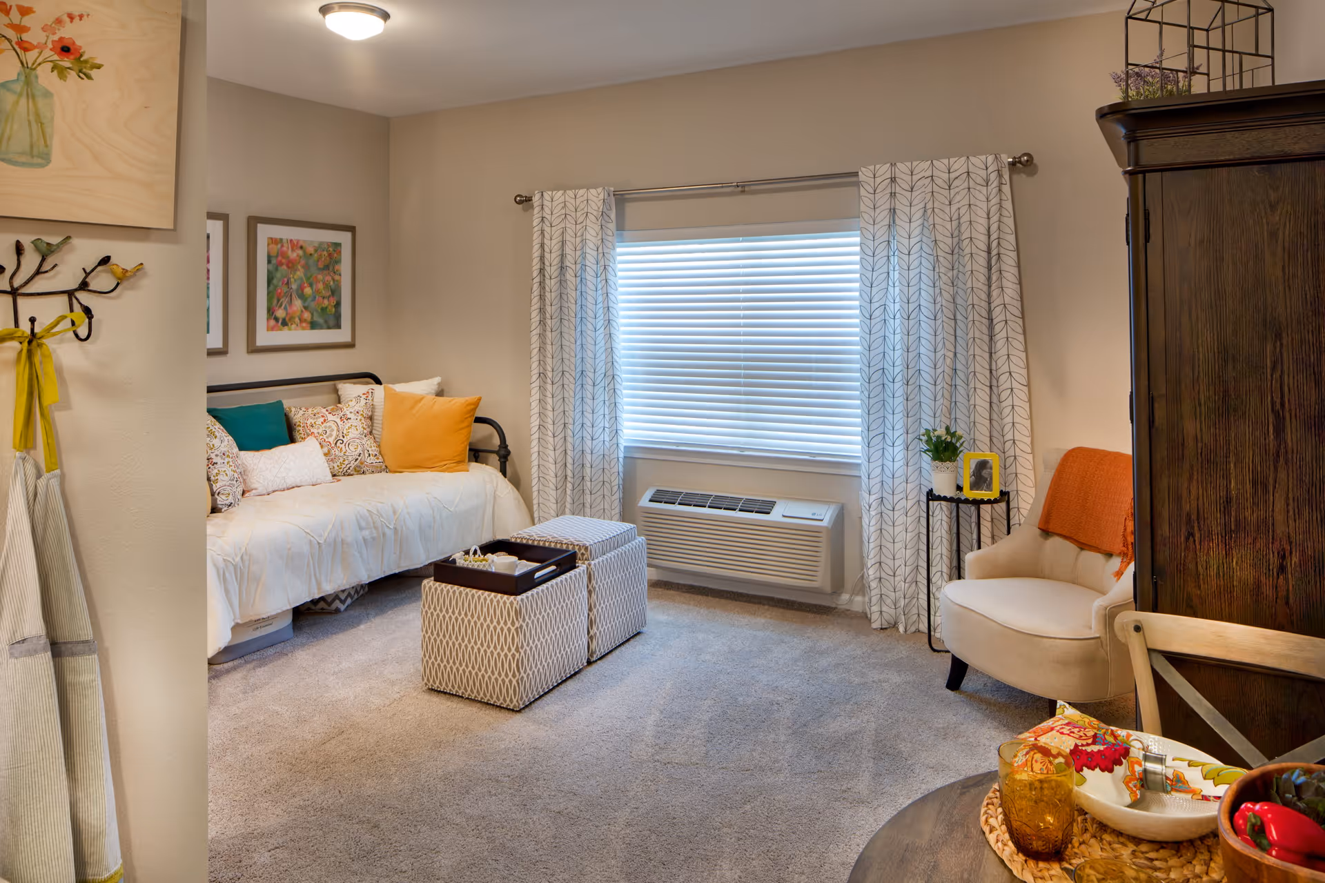 A cozy living room area with a daybed adorned with colorful pillows, two patterned ottomans, a beige armchair with an orange throw, a small round table with decorative items, a window with white blinds and patterned curtains, and a dark wooden cabinet.
