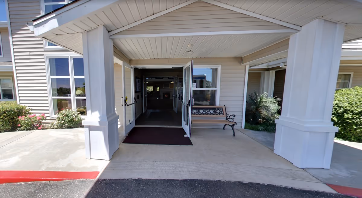 Entrance to a building with double glass doors open, a bench to the right, white columns supporting a covered porch, and some greenery and flowers on the sides.