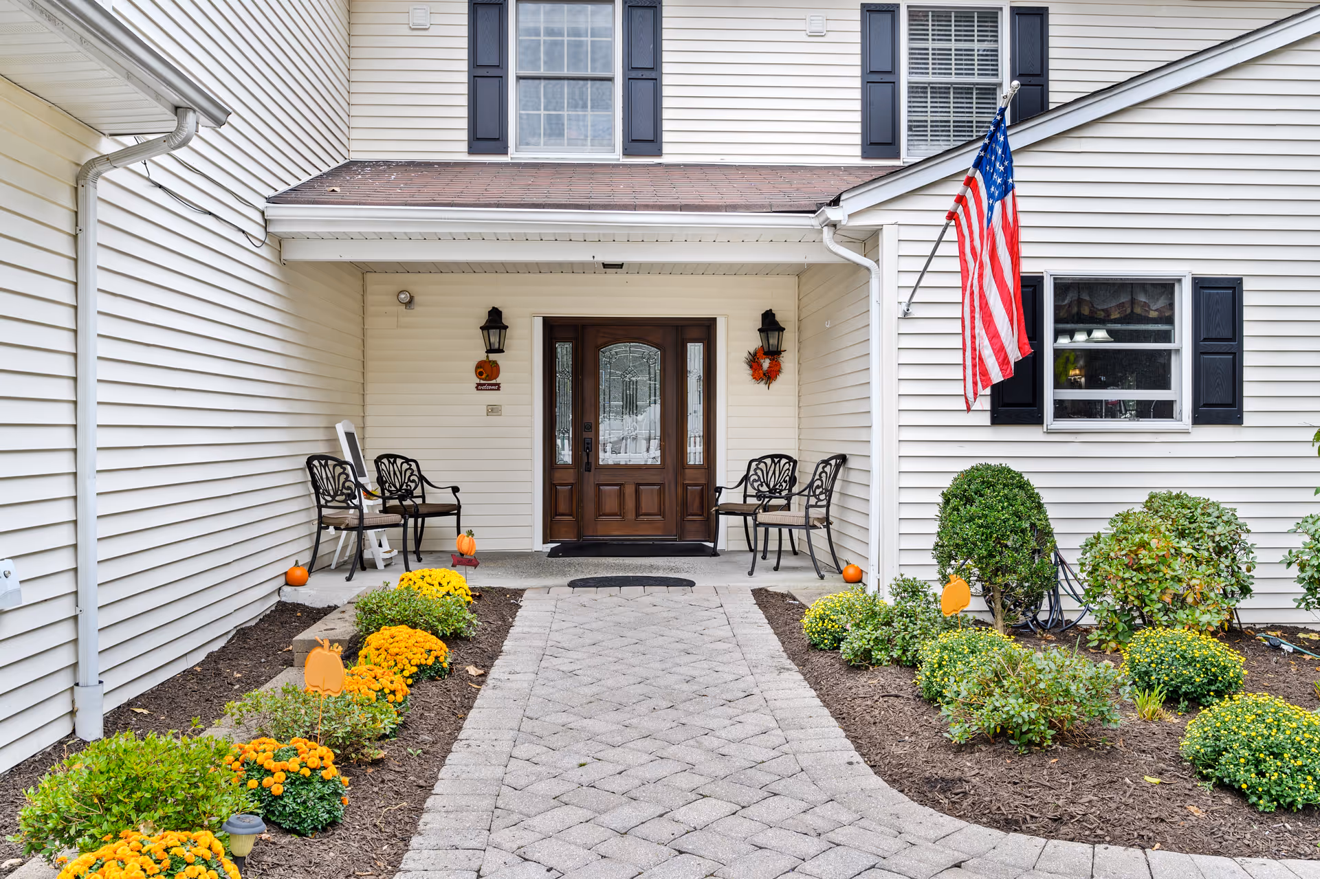 Front entrance of a memory care facility with walkway, seating, American flag, and blooming shrubs