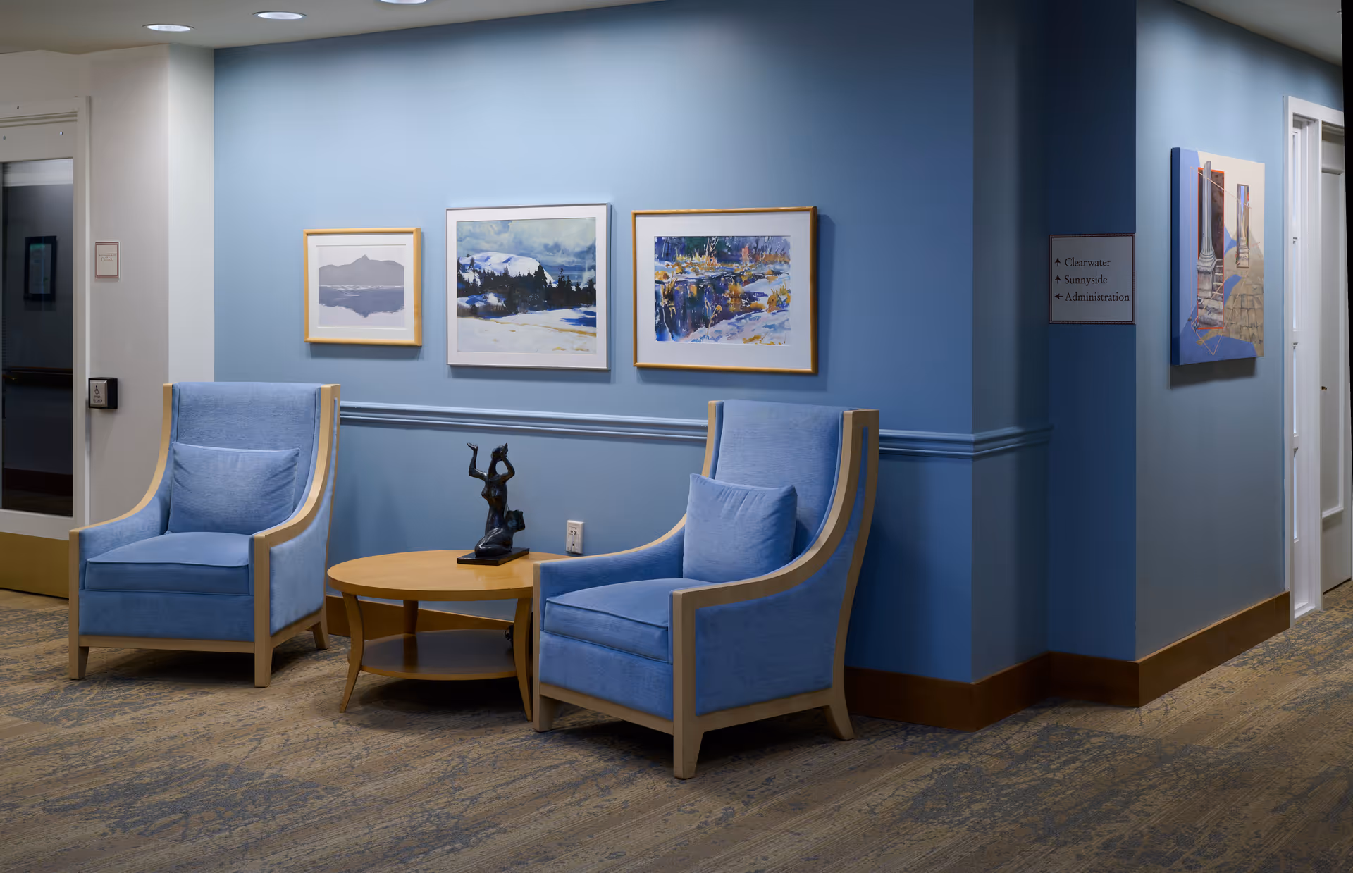 A cozy seating area in a senior living facility with two light blue armchairs and a round wooden coffee table between them. On the table is a small black sculpture. The walls are painted light blue and decorated with three framed landscape paintings. There is a hallway visible to the right with a sign indicating directions to Clearwater, Sunnyside, and Administration.