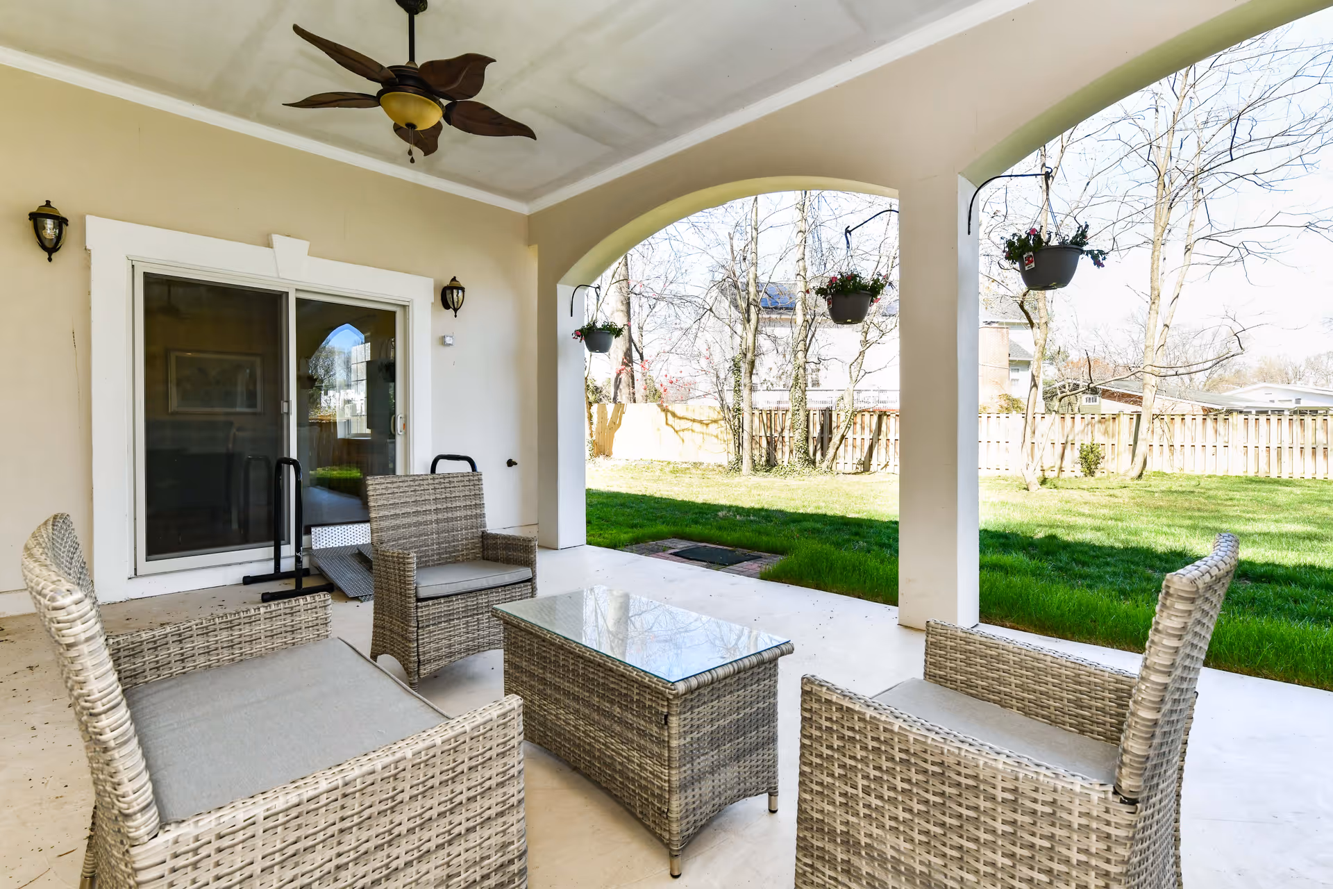 Covered outdoor patio area with wicker furniture including two chairs, a loveseat, and a glass-top coffee table. The patio has a ceiling fan with leaf-shaped blades and hanging flower pots. Beyond the patio is a grassy yard enclosed by a wooden fence with leafless trees visible.