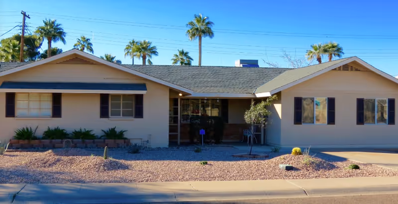 Single-story beige building with a dark shingled roof, several windows with dark shutters, and a small covered entrance. The front yard is landscaped with gravel, small plants, and a few palm trees in the background under a clear blue sky.