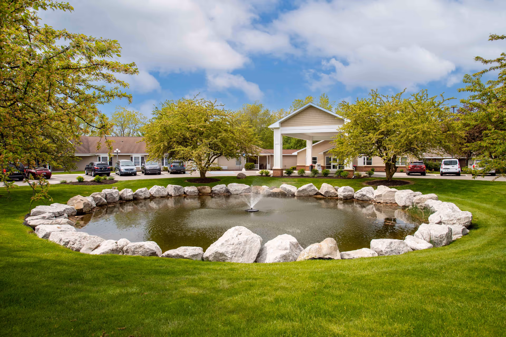Exterior view of Commonwealth Senior Living at East Paris featuring a stone-bordered pond and green lawn.