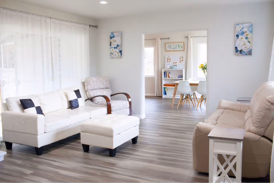 A bright lounge area with white furniture and a view of a dining space.