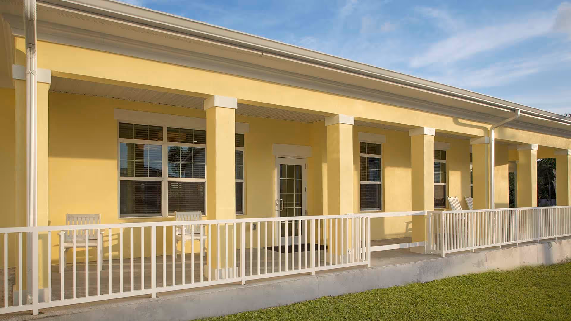 Yellow single-story building with a covered porch, white columns and railing, windows and chairs overlooking a grassy lawn.