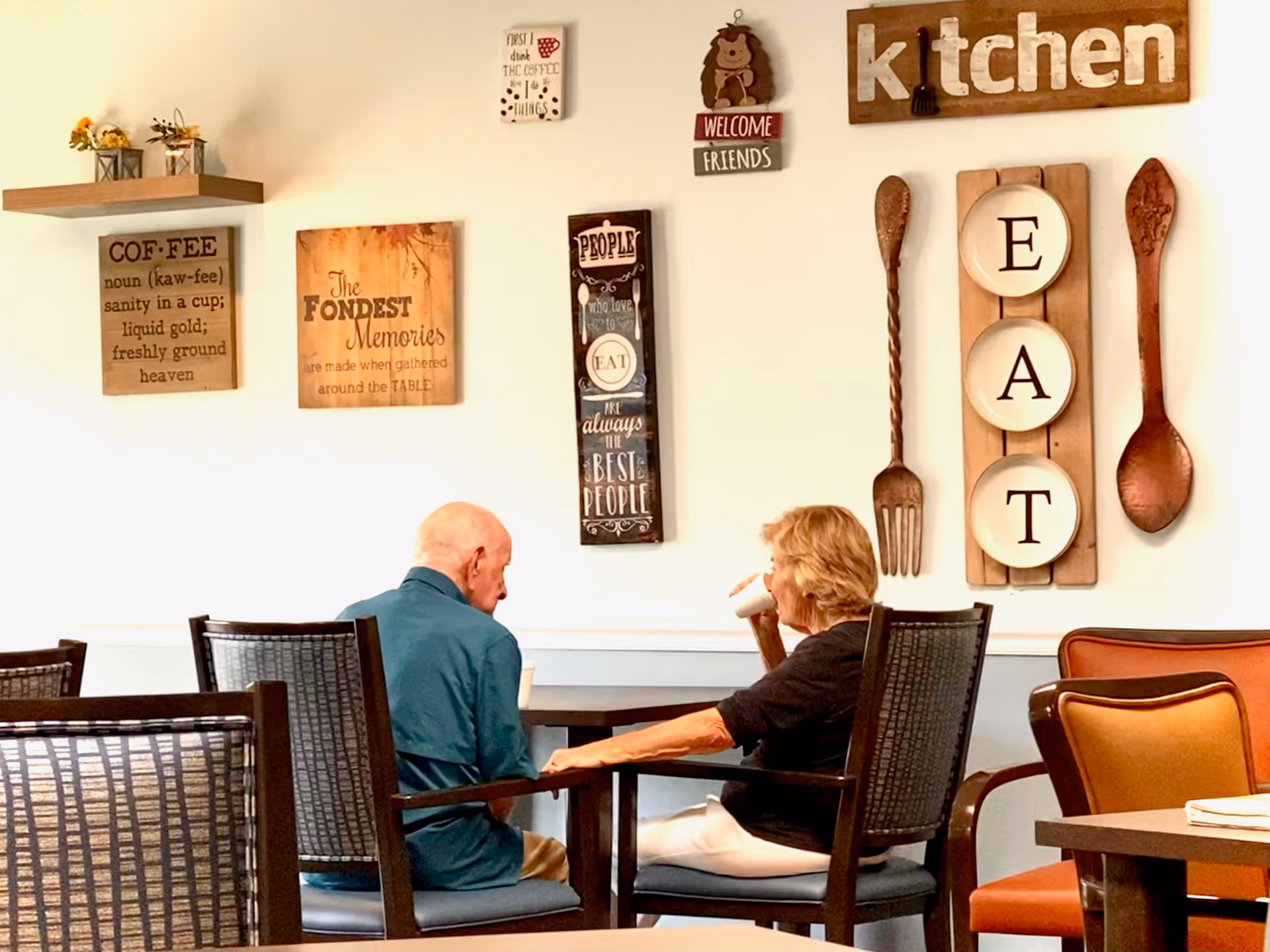 Two people sit at a table in a cozy dining area decorated with wooden kitchen signs and wall art.