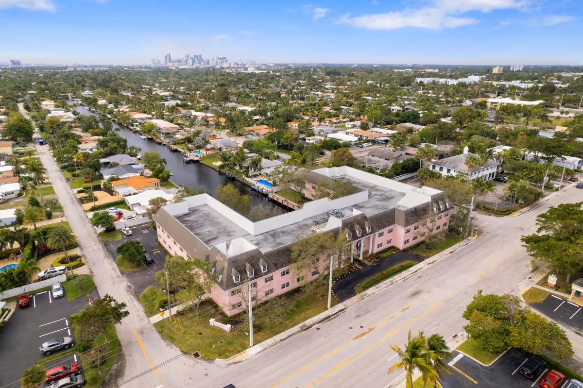 Aerial view of a pink assisted living building by a canal in a residential neighborhood with a distant city skyline.