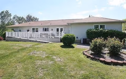 Single-story building with white siding and a brown roof, featuring a white railing ramp leading to the entrance. The building is surrounded by a well-maintained lawn with bushes and a small garden area under a partly cloudy sky.