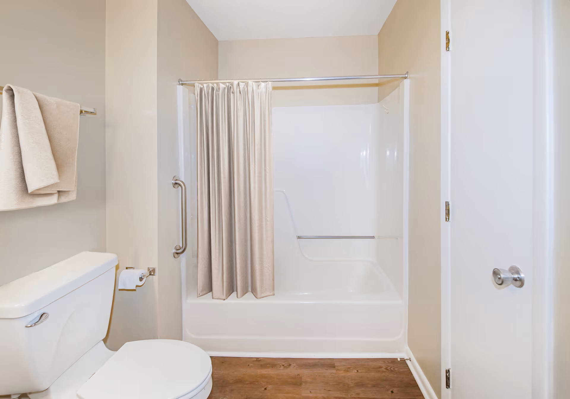A clean bathroom featuring a white toilet with a toilet paper holder and beige towels hanging on a towel rack. There is a white bathtub with a beige shower curtain partially drawn, and a metal grab bar mounted on the wall next to the tub. The walls are painted a light beige color and the floor has a wood-like finish.
