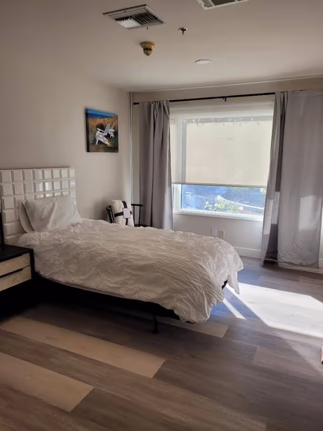 A bright bedroom with a single bed covered in a white comforter and pillow. There is a nightstand with drawers next to the bed. A window with gray curtains and a white roller shade allows natural light to fill the room. A small painting of white chairs on a beach hangs on the wall above the bed.