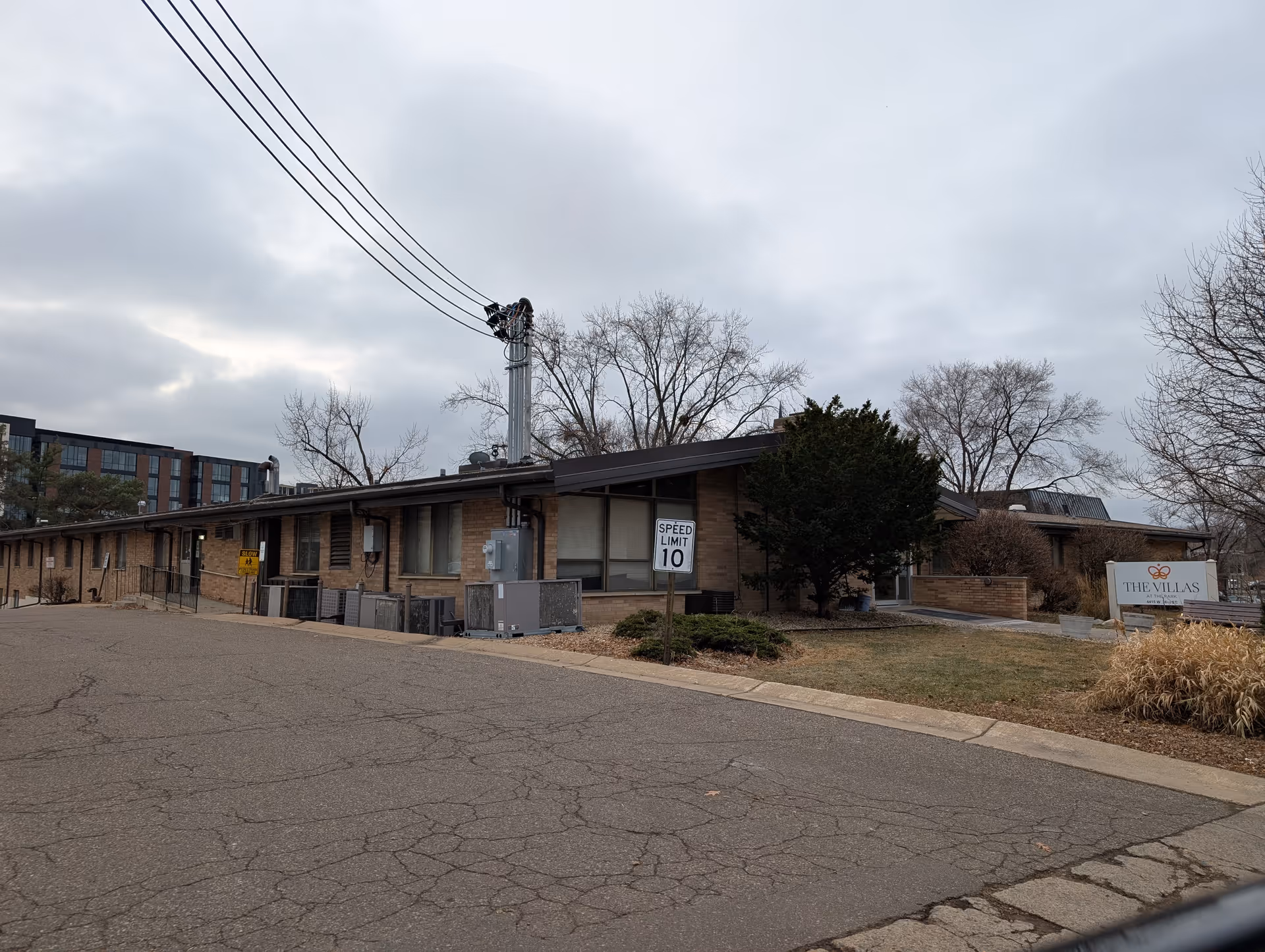Exterior view of a single-story brick building with a slanted roof, surrounded by leafless trees and a cloudy sky. There is a speed limit sign showing 10 mph and a sign that reads 'THE VILLAS' near the entrance.