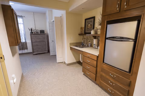 Interior view of a senior living facility showing a small kitchenette area with wooden cabinets, a mini refrigerator, a sink, and countertop. In the background, there is a carpeted room with a chest of drawers and a window with curtains.