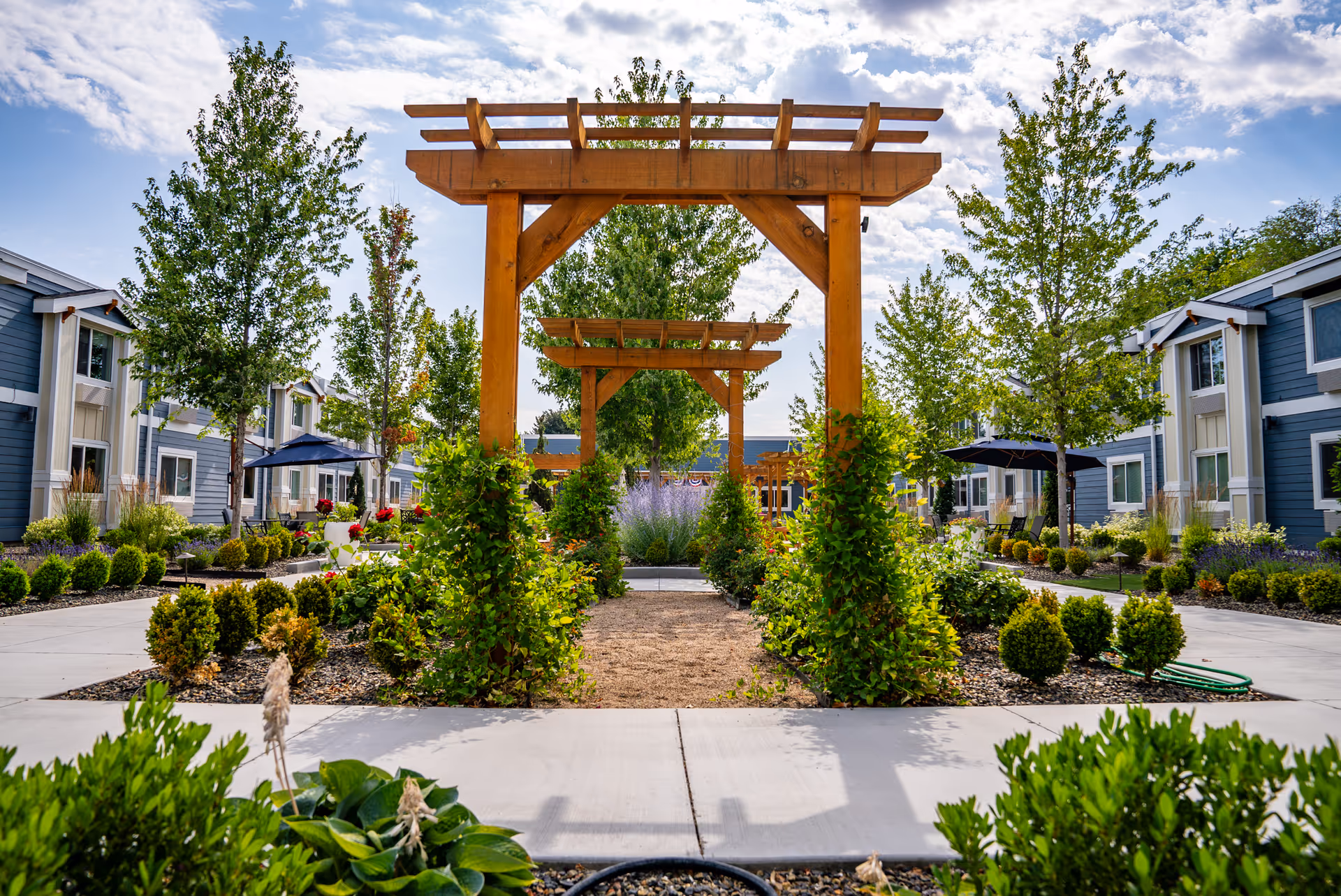 Outdoor garden area at Heatherwood Senior Living featuring wooden pergolas, green shrubs, trees, and a paved walkway. Blue sky with scattered clouds is visible above, and two-story blue and white buildings flank the garden on both sides.