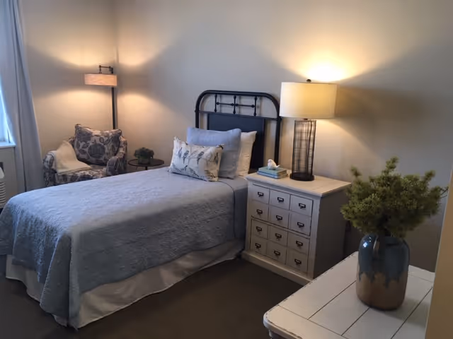 A tidy bedroom with a single bed dressed in blue linens, a nightstand with a lamp, an upholstered chair, and decorative plants.