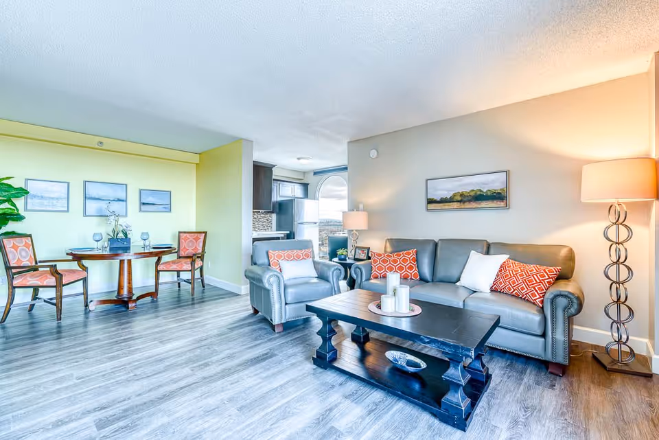 A bright and modern living room and dining area in a senior living facility. The living room features a gray leather sofa with orange and white patterned pillows, a matching armchair, a dark wooden coffee table with decorative candles, and a tall floor lamp with a circular design. The dining area has a round wooden table with two chairs upholstered in orange patterned fabric, three framed landscape pictures on a yellow wall, and a potted plant. The kitchen is partially visible in the background with dark cabinets and a stainless steel refrigerator.