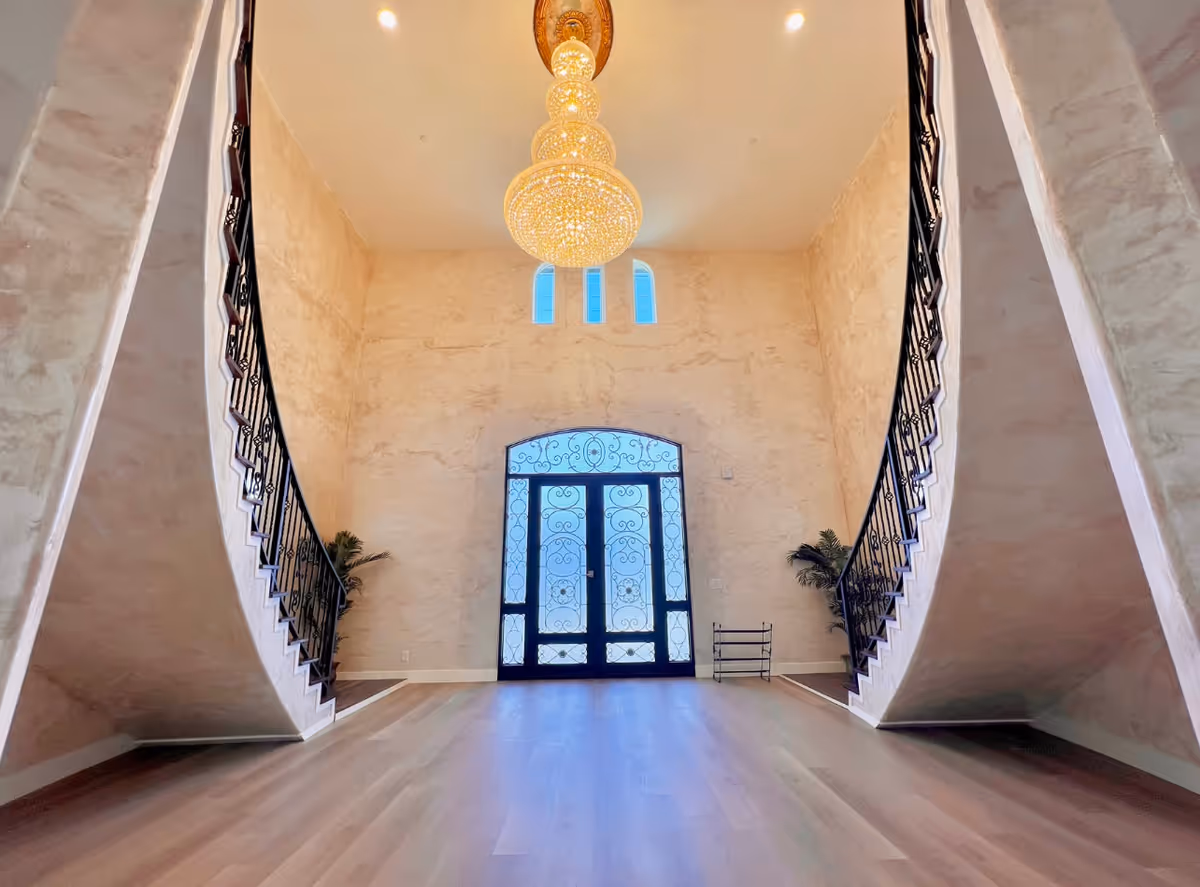 Spacious entrance hall with a large ornate chandelier hanging from the ceiling, two symmetrical curved staircases with black wrought iron railings on either side, textured beige walls, and a decorative glass double door with intricate designs. There are two small plants placed near the door.