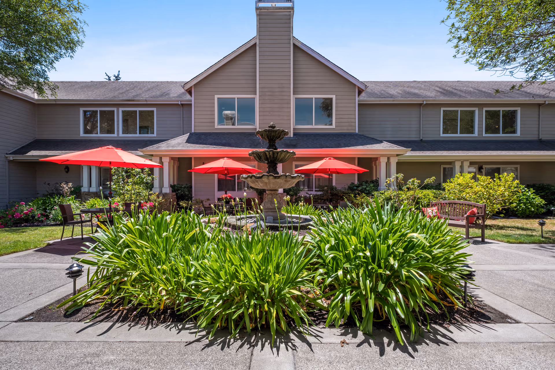 Outdoor courtyard area of a senior living facility with a central multi-tiered fountain surrounded by green plants. There are red umbrellas shading seating areas with chairs and benches. The building in the background is two stories with beige siding and multiple windows.