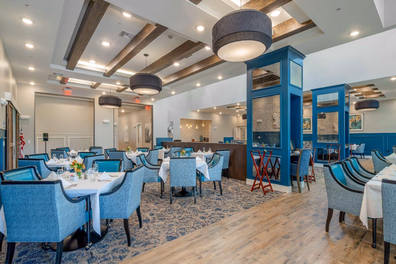 A spacious and elegant dining room with multiple tables covered in white tablecloths, each set with glassware, napkins, and small floral centerpieces. The room features blue upholstered chairs, wooden ceiling beams, large round pendant lights, and blue decorative columns with mirrors. The floor is a combination of patterned carpet and wood.